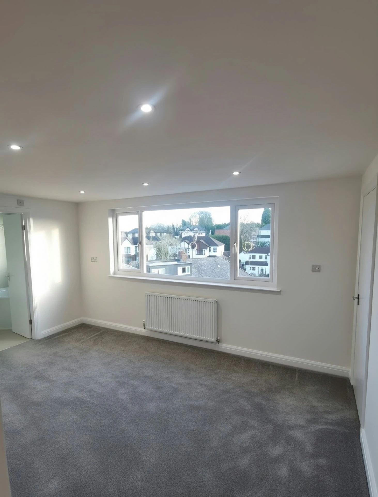 Finished carpeted room in the Prestwich loft conversion with a wide window, radiator and recessed ceiling lights