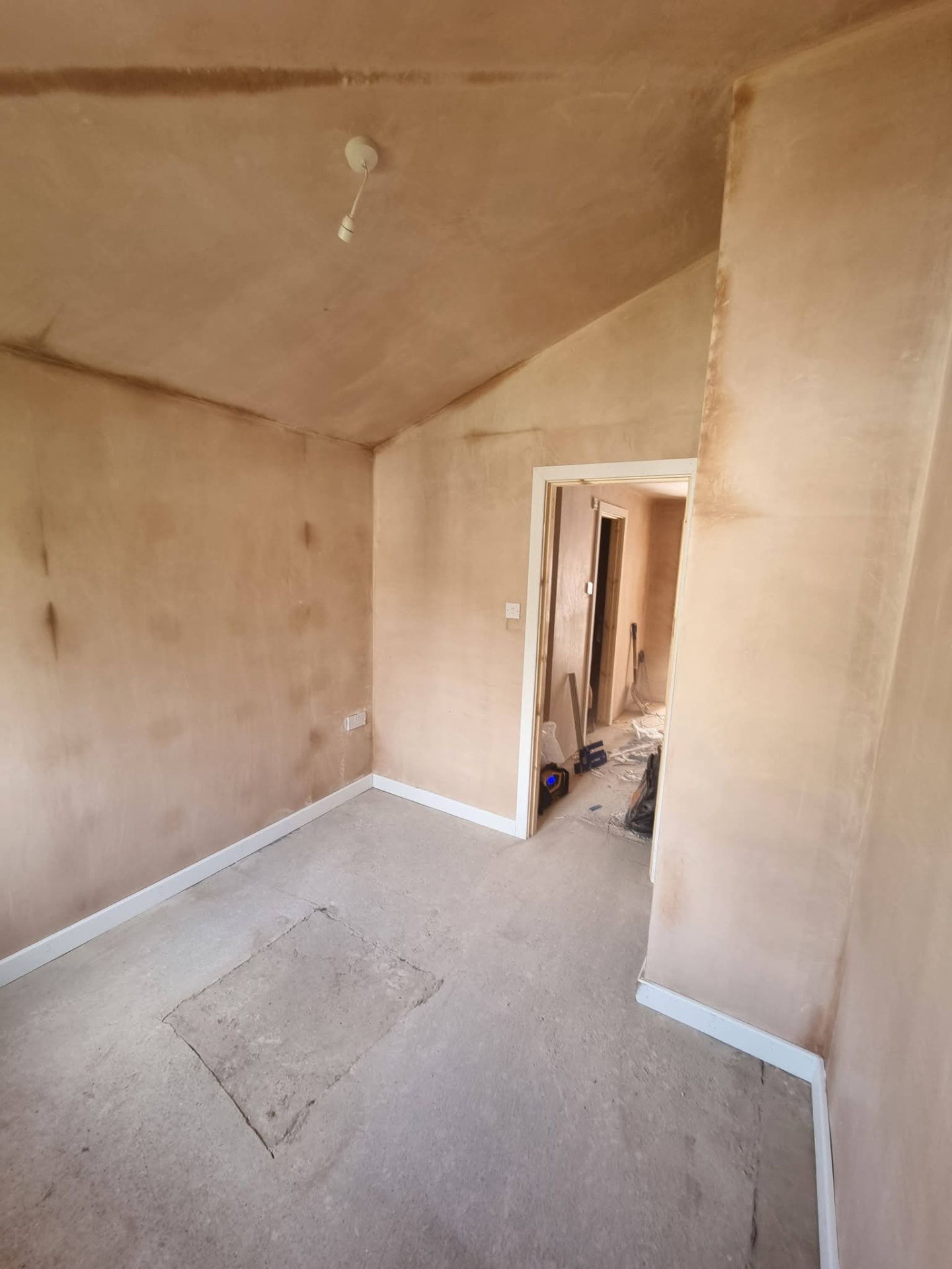 Newly plastered room with sloping ceiling, white skirting and unfinished concrete floor